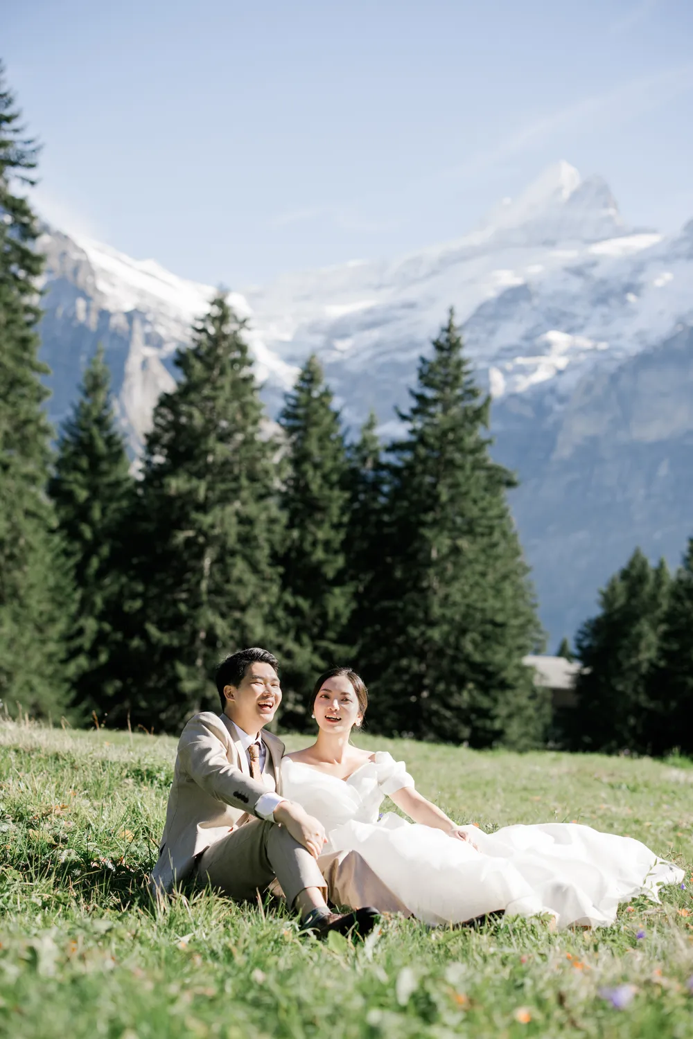 Couple sitting on a sun-drenched meadow in Grindelwald with the vast Swiss Alps and peaks rising behind them.