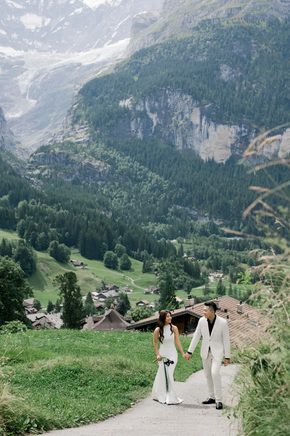 Romantic portrait of a **Malaysian couple** holding hands in **Grindelwald** with a traditional yellow Swiss hiking sign and chalet.